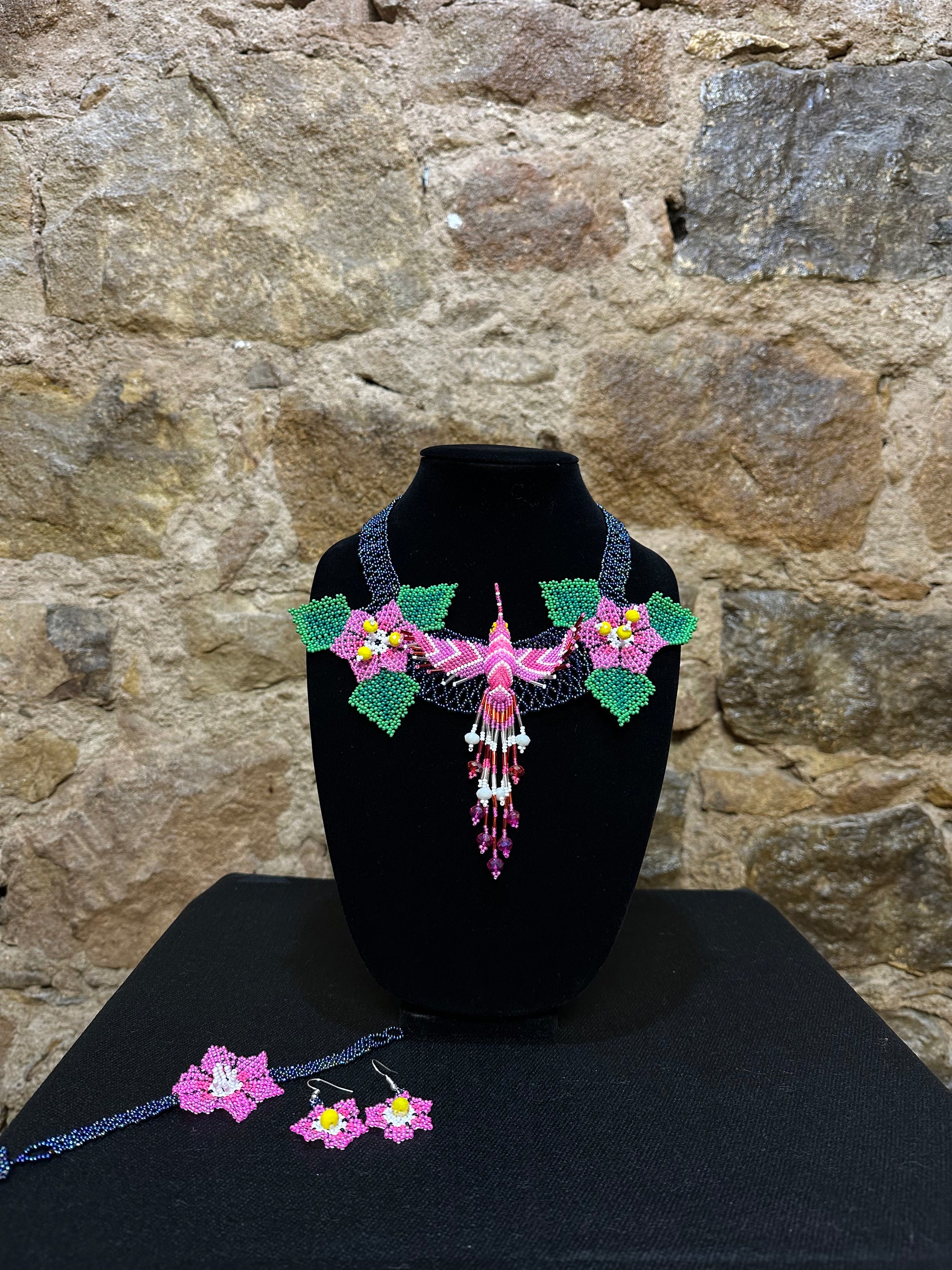 This vibrant hummingbird necklace and earring set are sure to bring a touch of cheerful color to any look. Expertly crafted, each set is made up of three-dimensional beading of flower set among three leaves with a 6" central hummingbird motif. Allowing you to wear a bit of nature's beauty with you, this set is the perfect accessory for any warm weather ensemble!