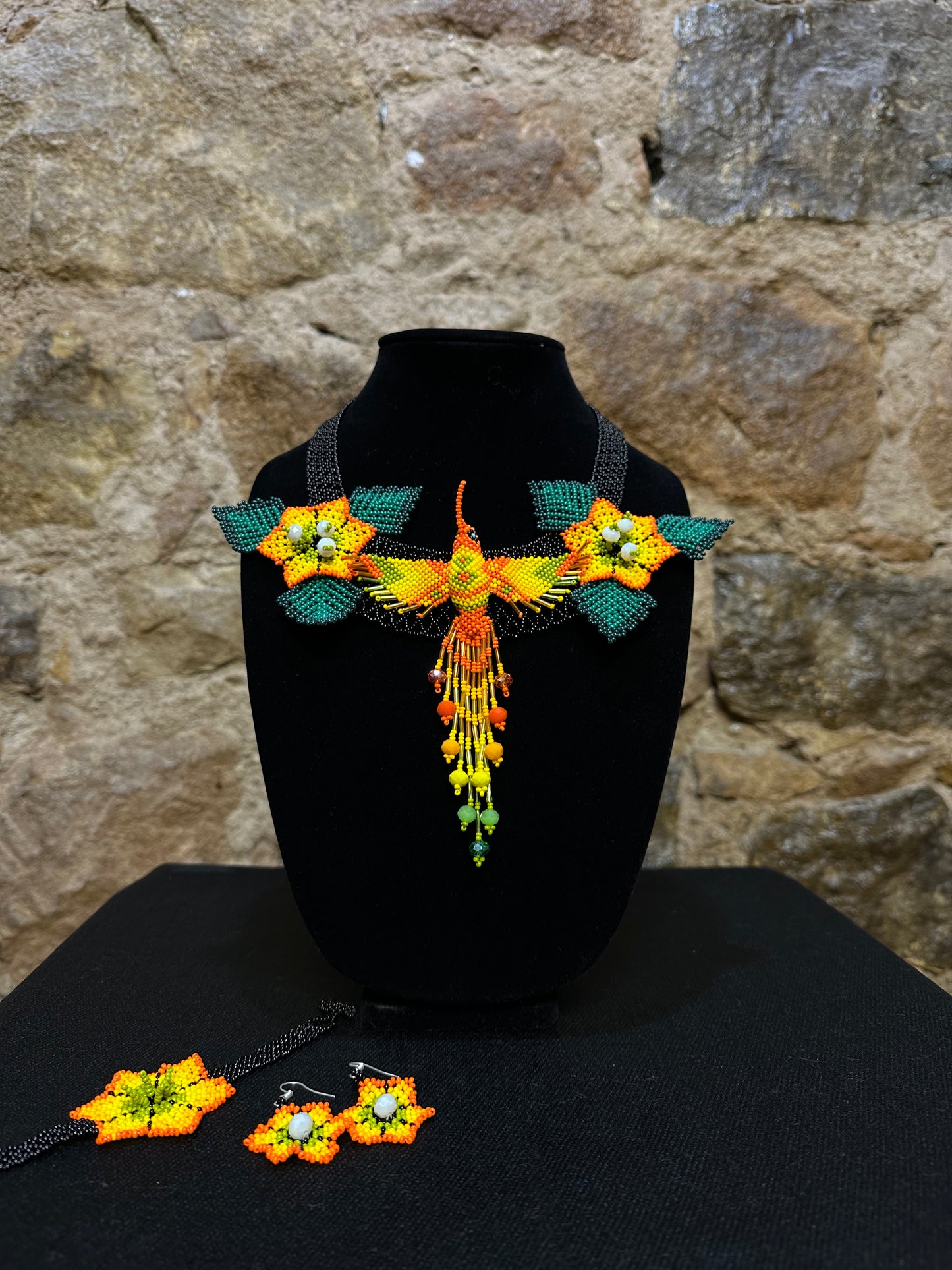 This vibrant hummingbird necklace and earring set are sure to bring a touch of cheerful color to any look. Expertly crafted, each set is made up of three-dimensional beading of flower set among three leaves with a 6" central hummingbird motif. Allowing you to wear a bit of nature's beauty with you, this set is the perfect accessory for any warm weather ensemble!