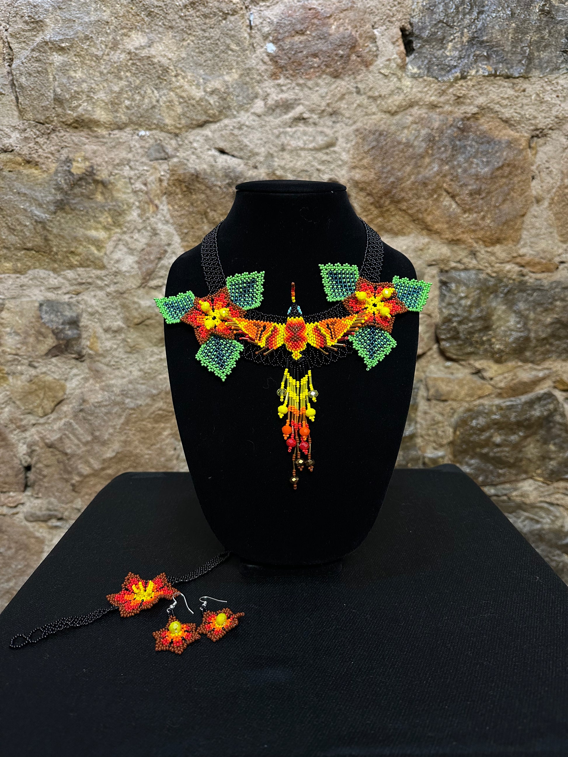 This vibrant hummingbird necklace and earring set are sure to bring a touch of cheerful color to any look. Expertly crafted, each set is made up of three-dimensional beading of flower set among three leaves with a 6" central hummingbird motif. Allowing you to wear a bit of nature's beauty with you, this set is the perfect accessory for any warm weather ensemble!