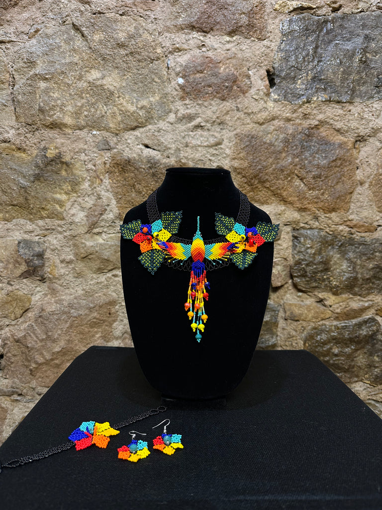 This vibrant hummingbird necklace and earring set are sure to bring a touch of cheerful color to any look. Expertly crafted, each set is made up of three-dimensional beading of flower set among three leaves with a 6